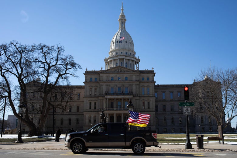 A car with a U.S. flag drives in front of the Michigan State Capitol.