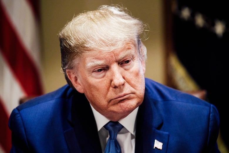 Donald Trump in a room with flags behind him, with a neutral expression.