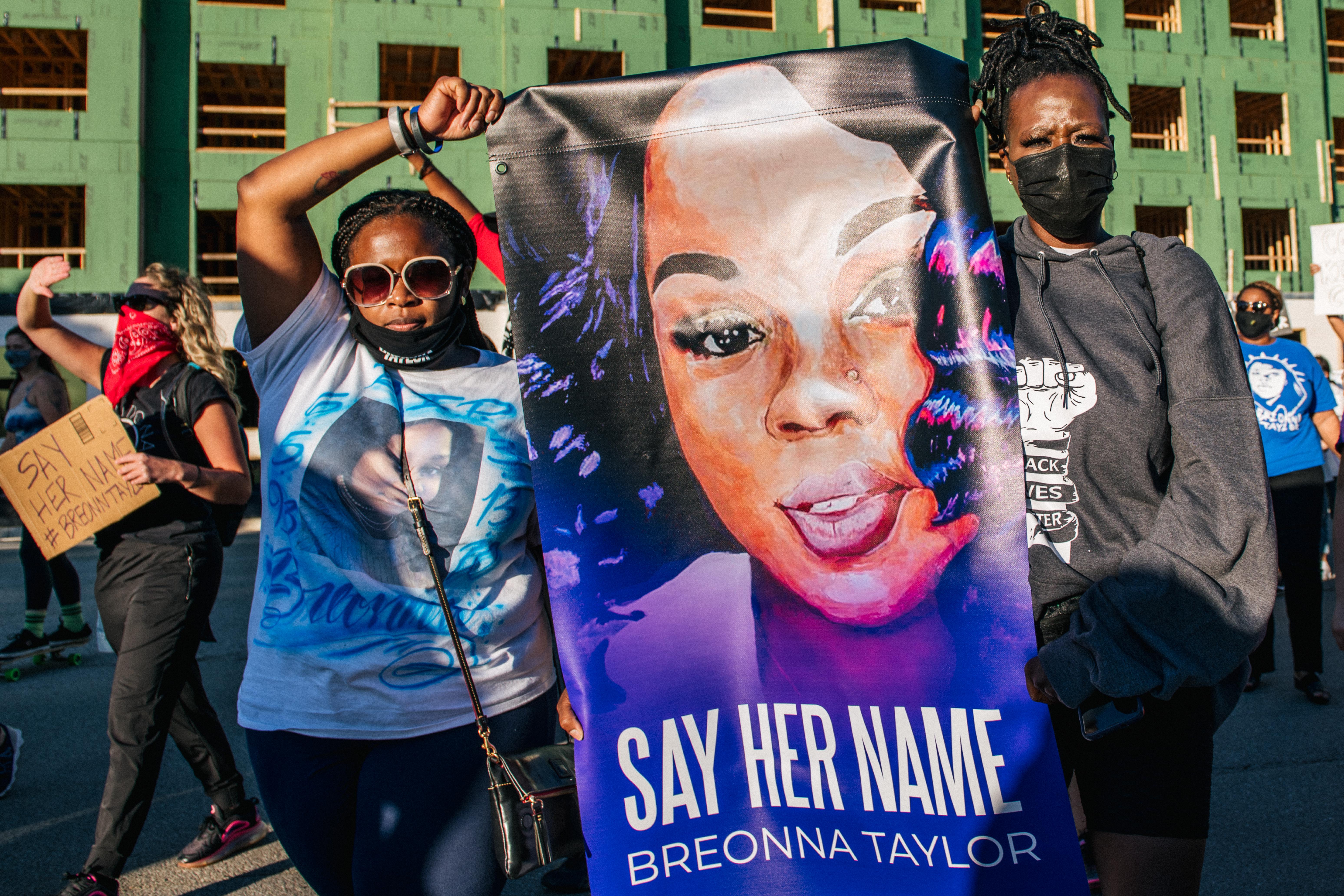 Two women at the front of a march hold a sign featuring an illustration of Breonna Taylor's face and the words "Say her name, Breonna Taylor."