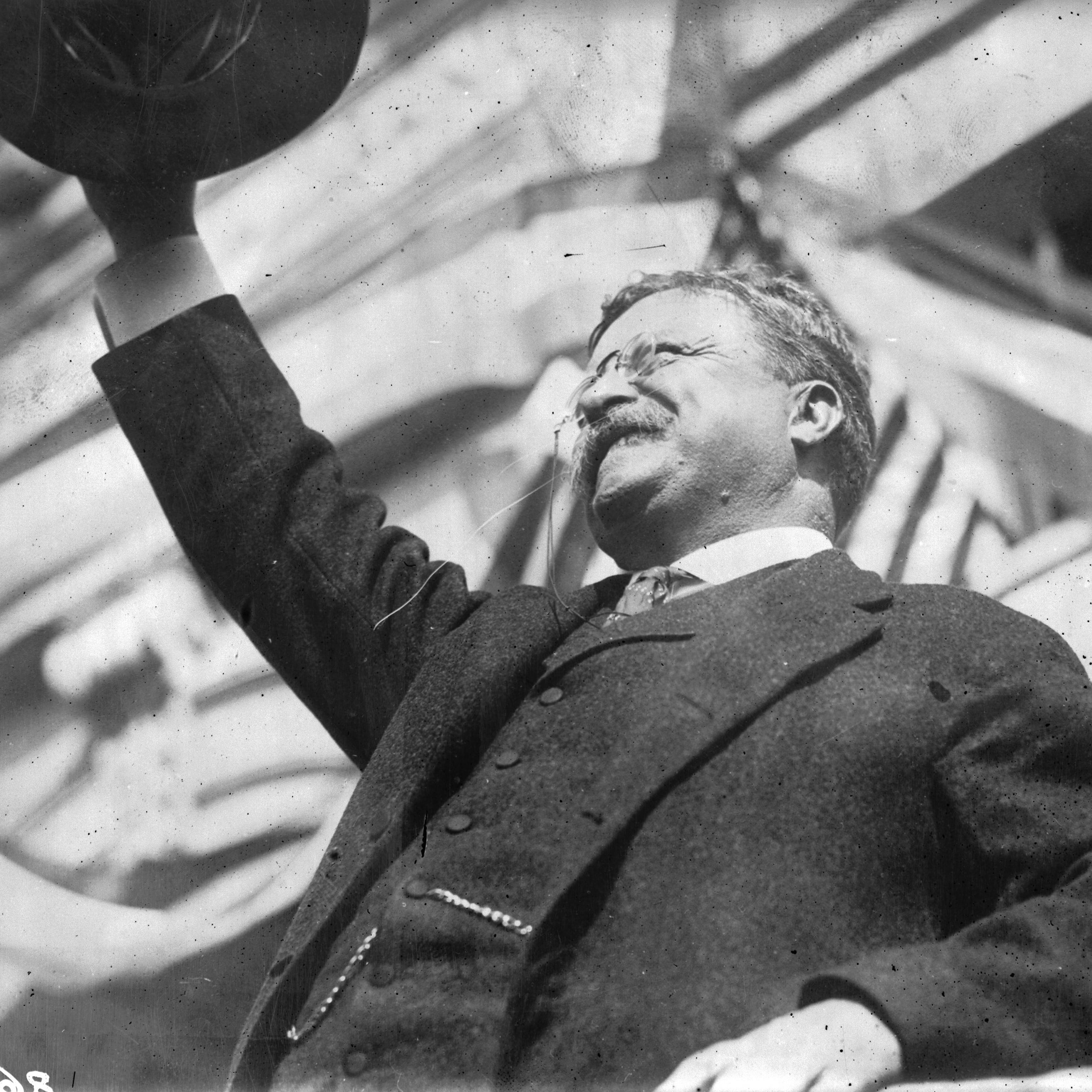 TR from below, smiling up at a crowd, raising his hat in the air