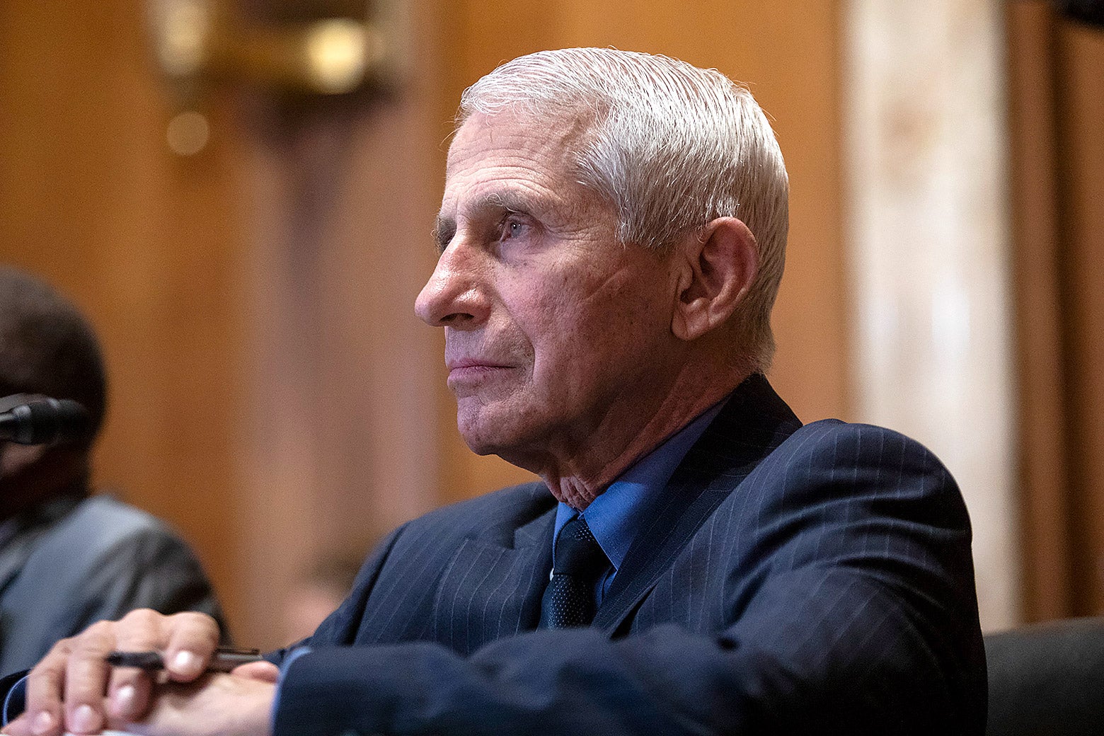 Dr. Anthony Fauci sitting calmly at a table with his hands folded and a pen between his fingers.
