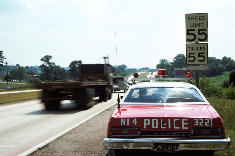 A red police car from the 1970s sits on the side of the highway.