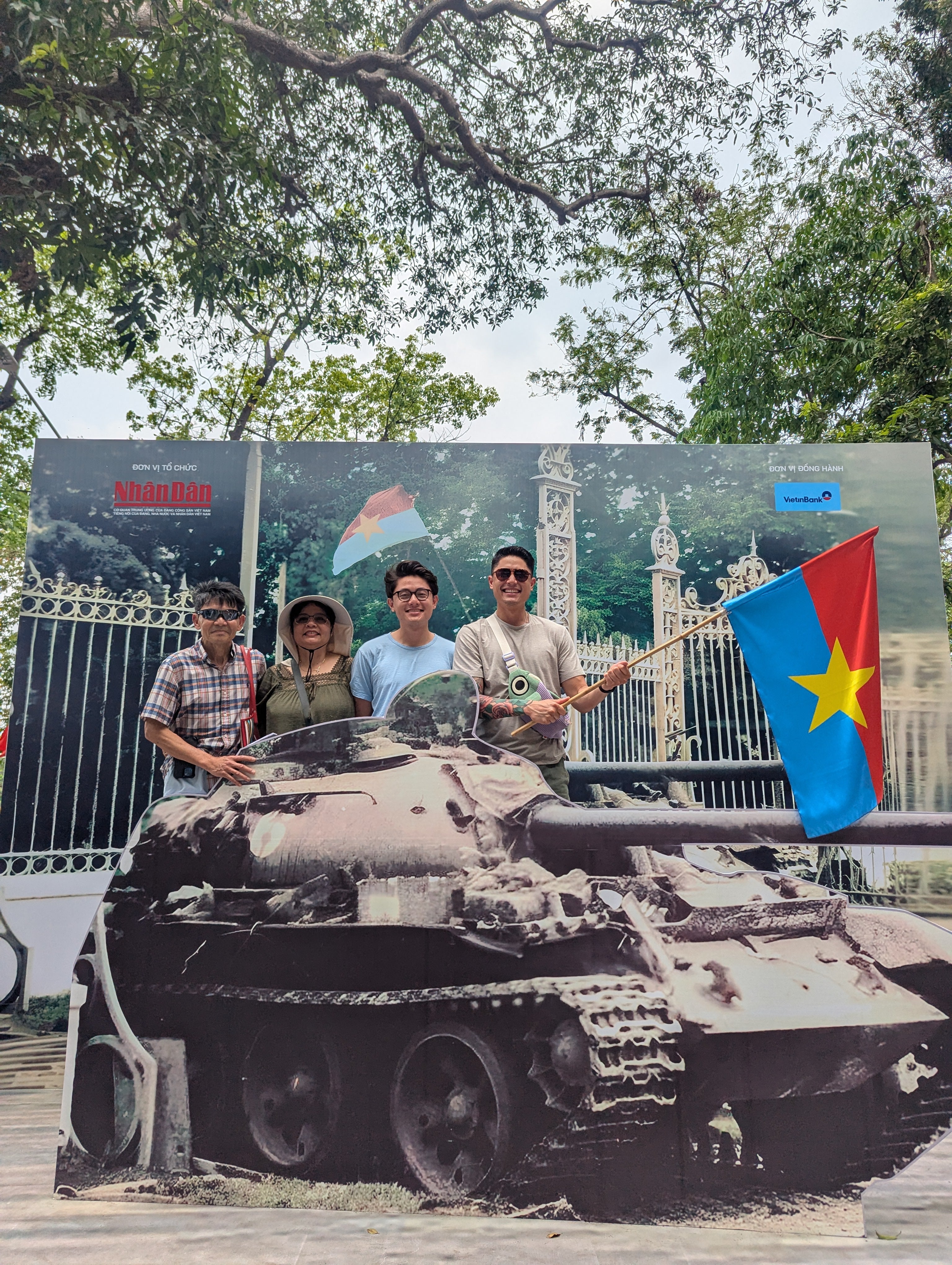 A family smiles and poses behind a cardboard cutout of a tank. One of the two adult sons holds a blue-and-red flag with a yellow star.