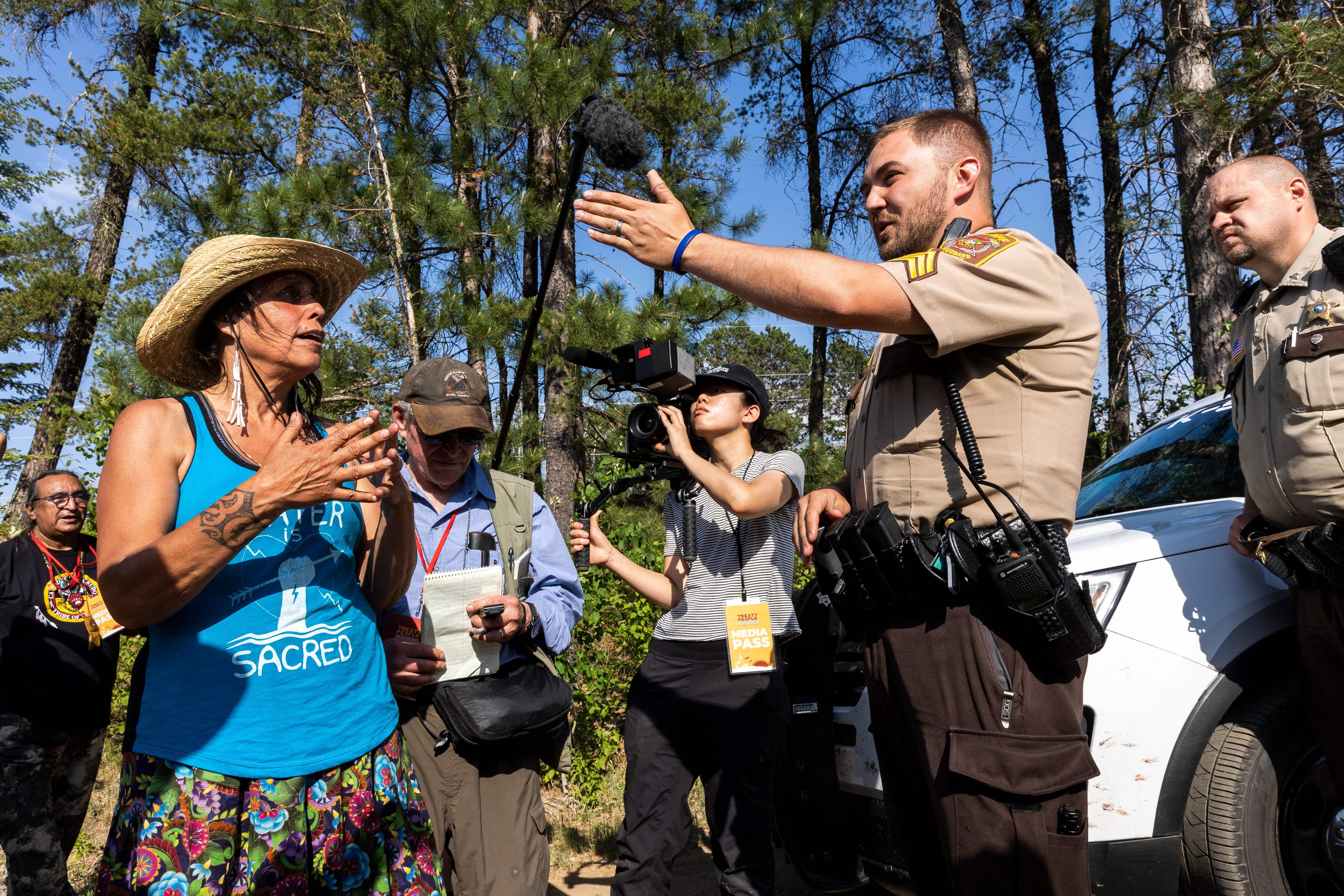 Winona LaDuke, wearing a blue tank top and tan straw hat, talks to the Hubbard County Sheriff in uniform as LaDuke's fellow protestors and media look on.