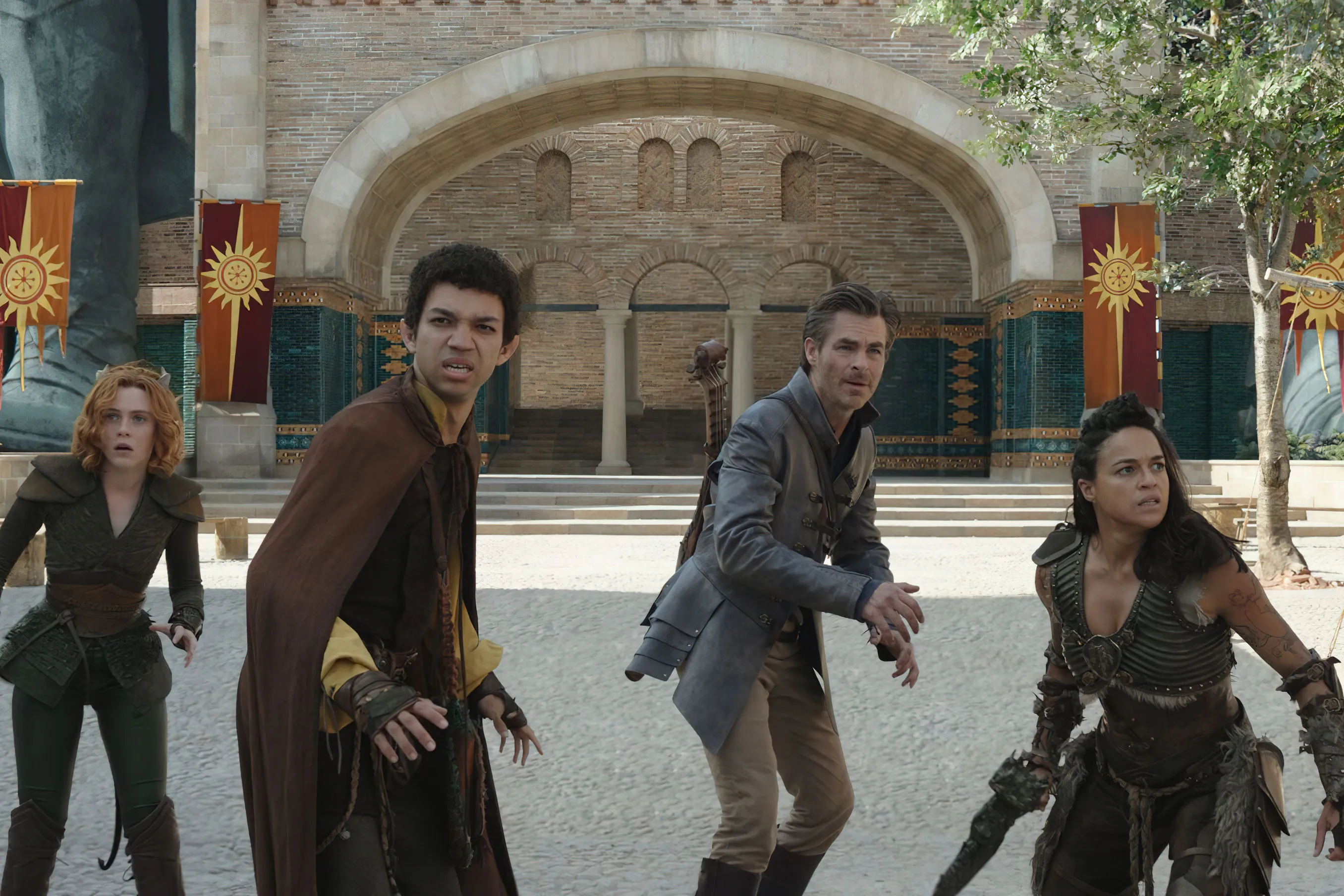 In a cobblestoned medieval-looking courtyard, four people stand in a line looking ahead in fear, ready to attack. From left to right: a female druid, a male half-elf sorcerer, a male bard, and a female barbarian.