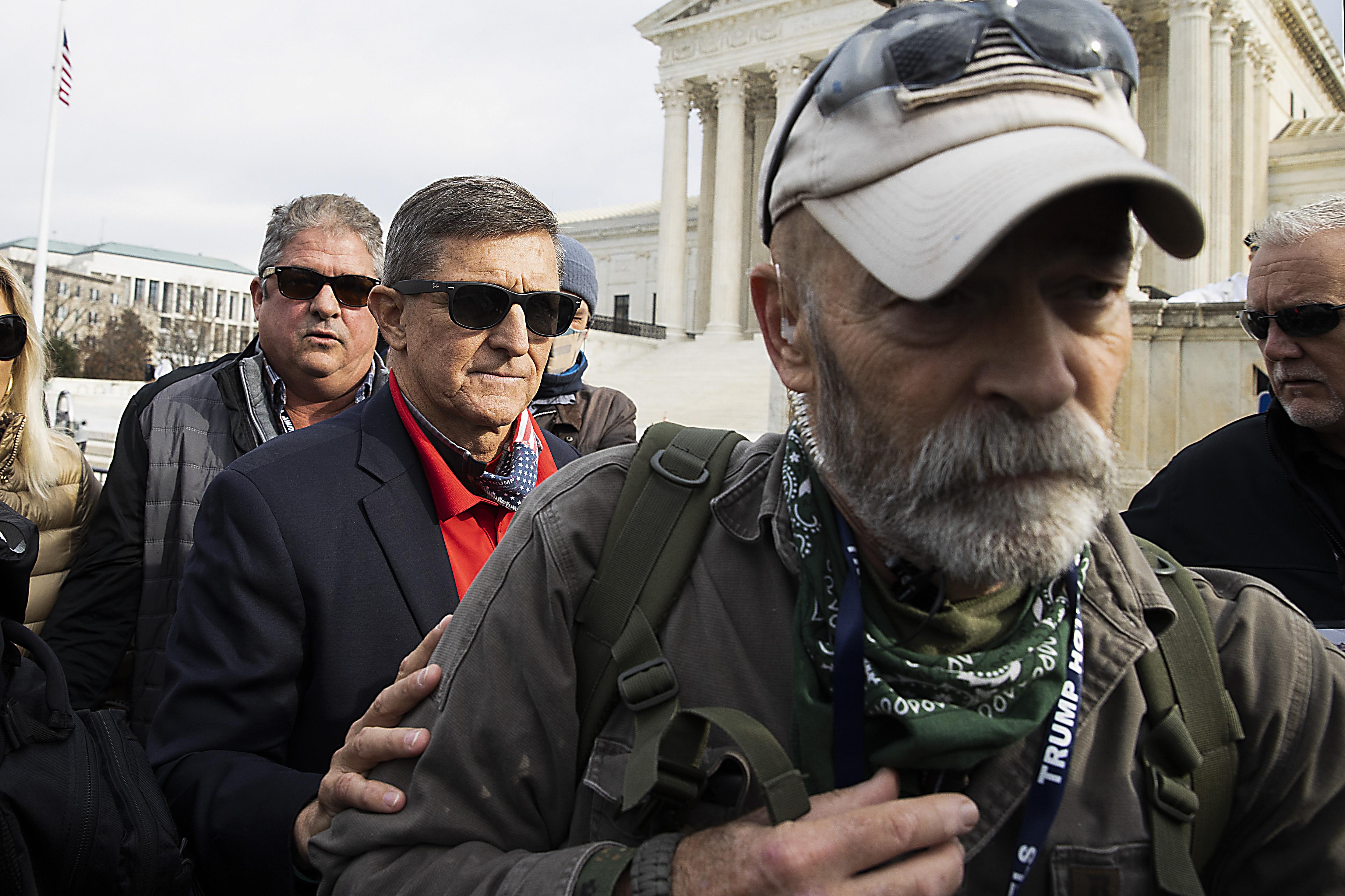 Michael Flynn standing in a group of protesters. Flynn wears sunglasses and puts his hand on the arm of a man in front of him who is wearing a Trump lanyard.