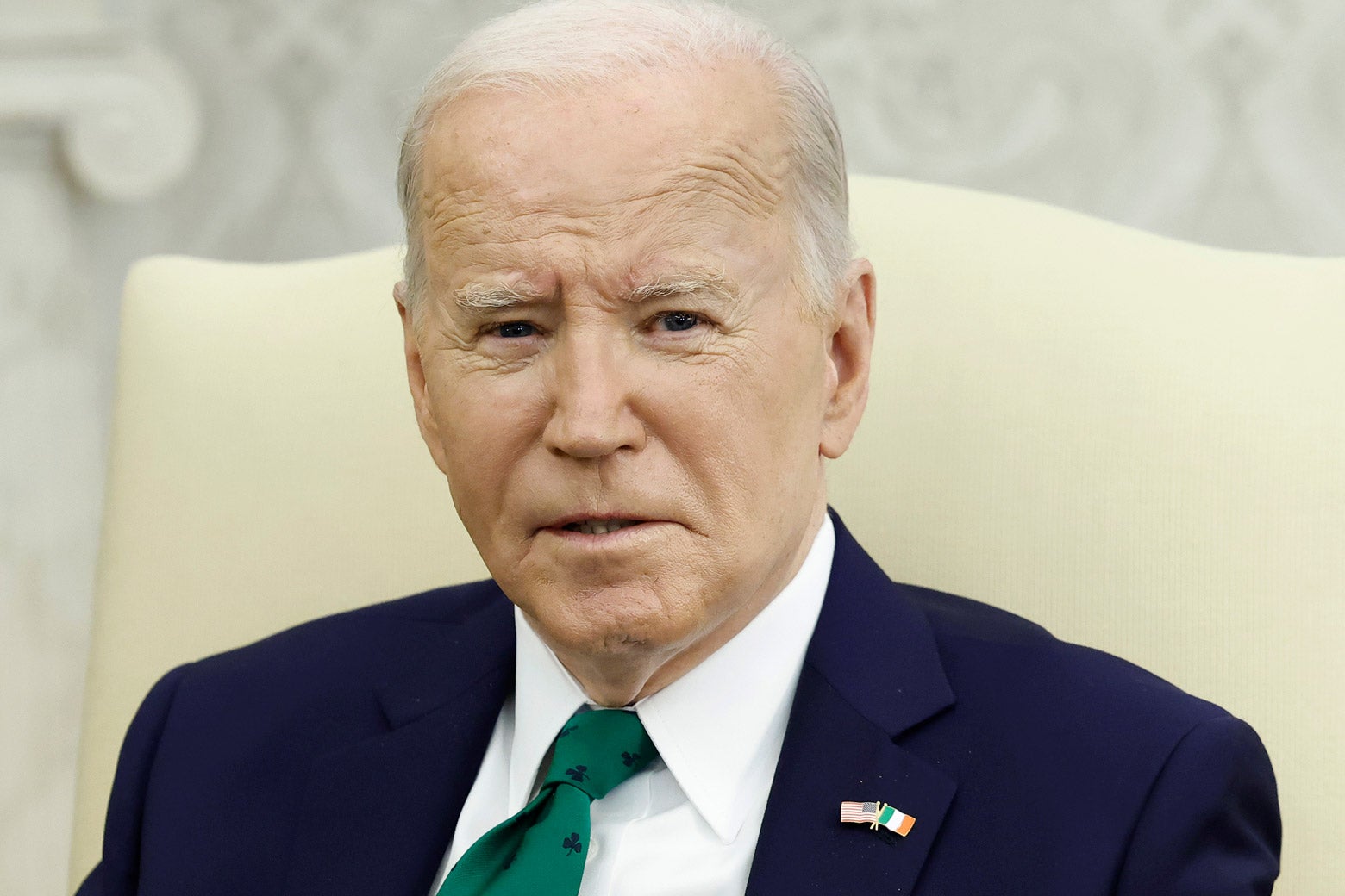 Biden seeming to grimace wearing a blue suit and sitting on a peach chair.