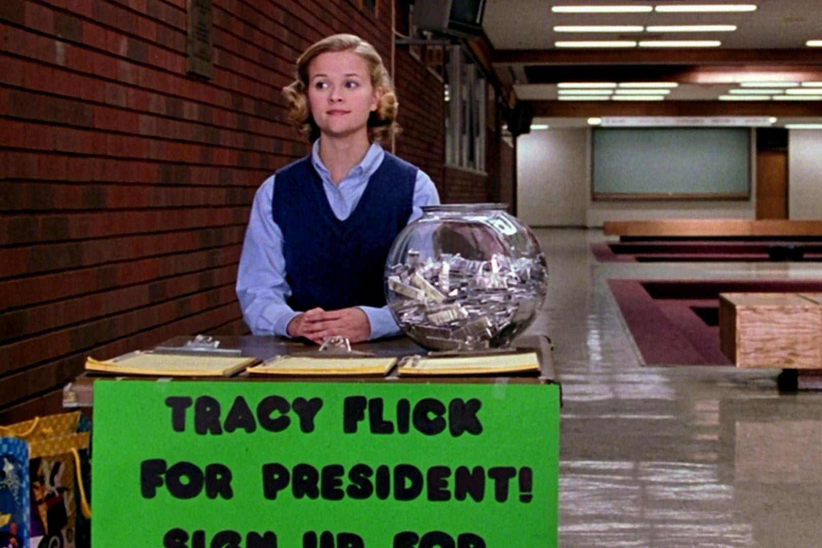 A young Reese Witherspoon sits as a student at a table in a High School hallway with a sign on it that says "Tracy Flick for president!" and some sign up sheets on the table. 