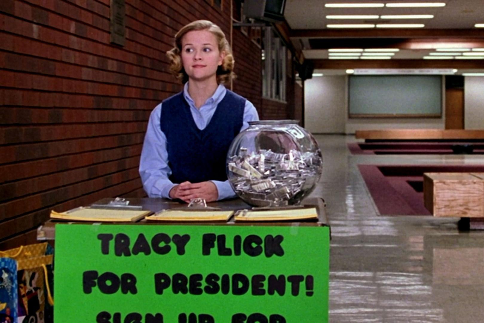 A young Reese Witherspoon sits as a student at a table in a High School hallway with a sign on it that says "Tracy Flick for president!" and some sign up sheets on the table.