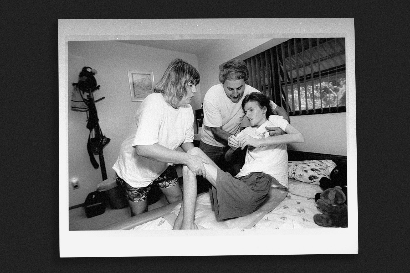A mother and father lower a very frail daughter into a bed in an archival black-and-white photo.