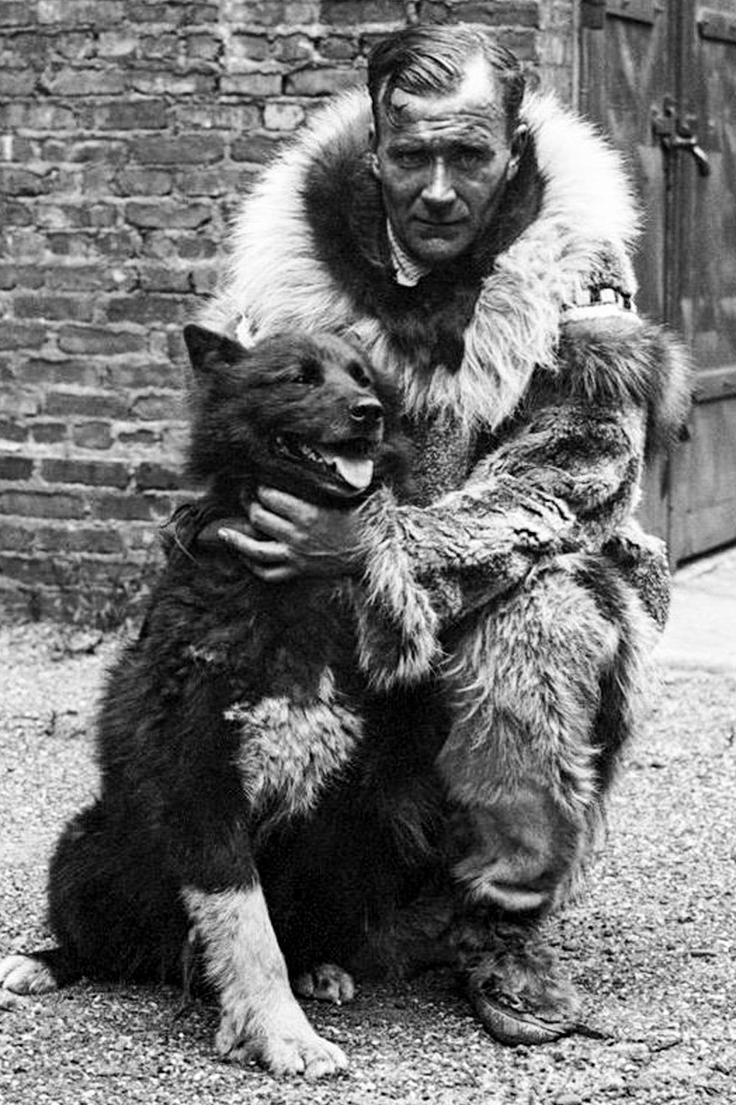 A man in a large fur jacket crouches next to a black and white dog.
