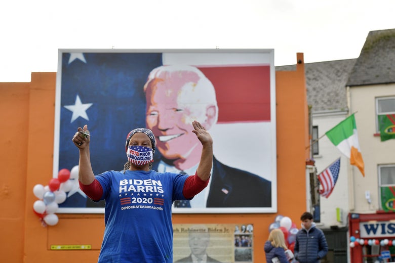 A Black woman wearing a Biden-Harris mask cheers in front of a mural with a painting of Joe Biden.