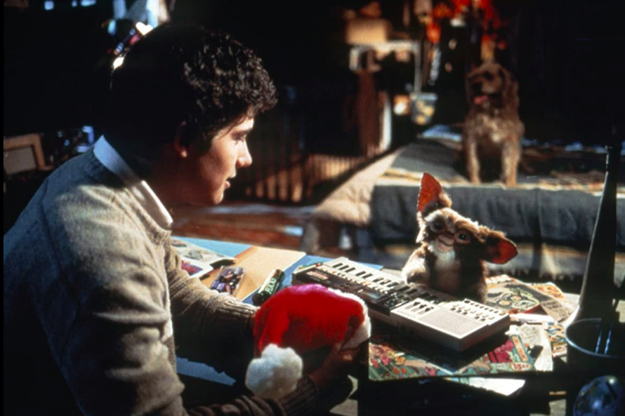 A small creature with big eyes and pointy ears sits at a keyboard in front of Zach Galligan, who holds a Santa hat.