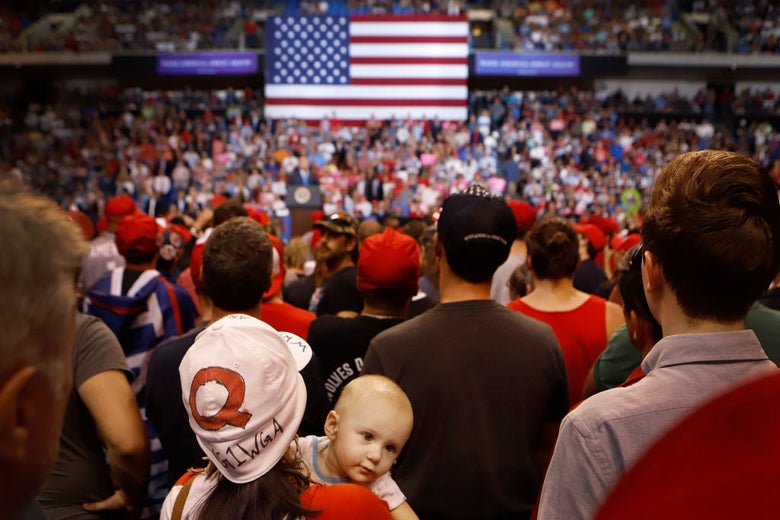 The woman in the Q hat holds a baby who is facing toward the camera. Trump can be seen in the distance speaking in front of an American flag.