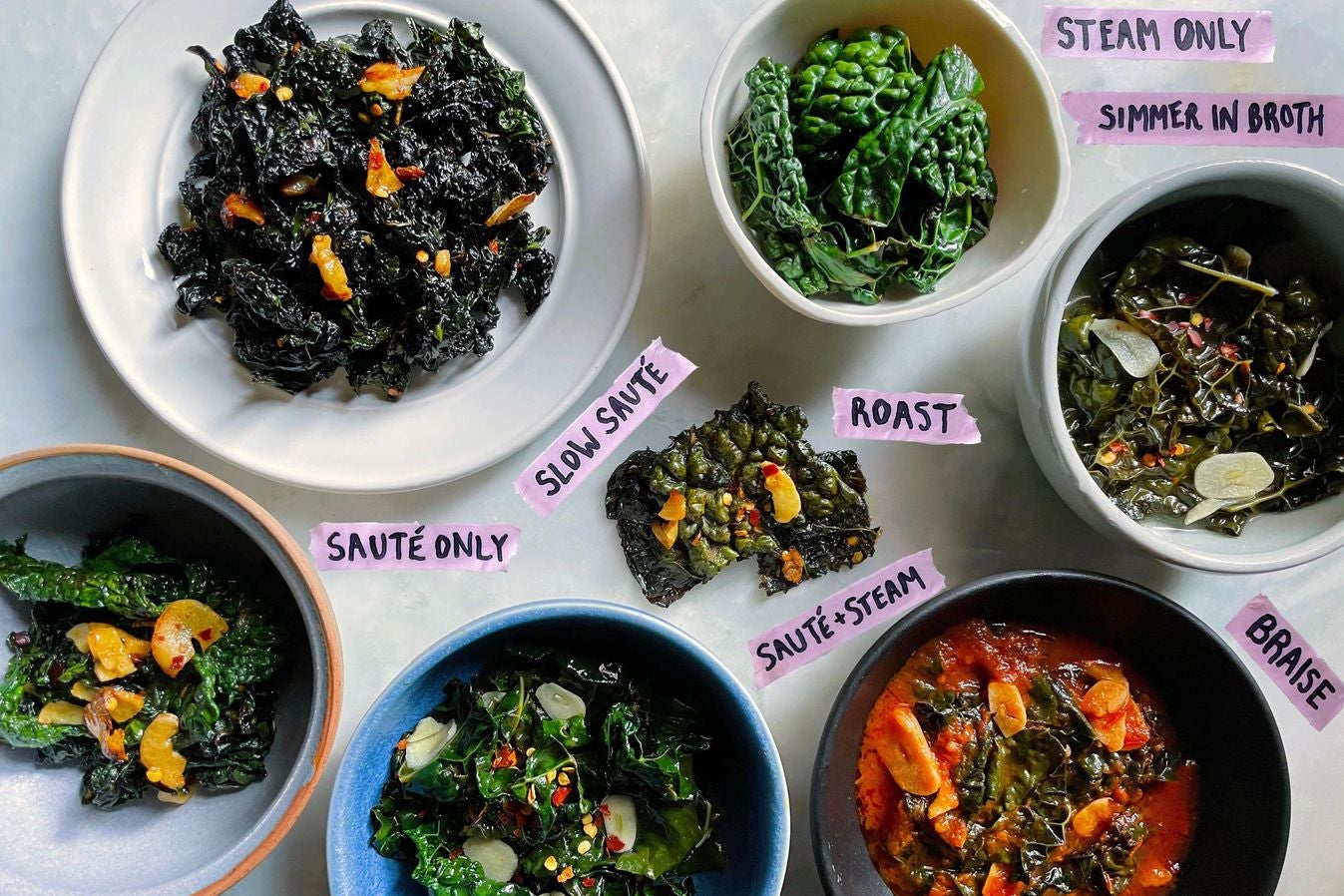 Seven different preparations of kale labeled sauté only, slow sauté, sauté and steam, roast, simmer in broth, steam only, and braise. All but the bowl of steam-only kale are topped with sliced garlic and red pepper flakes.