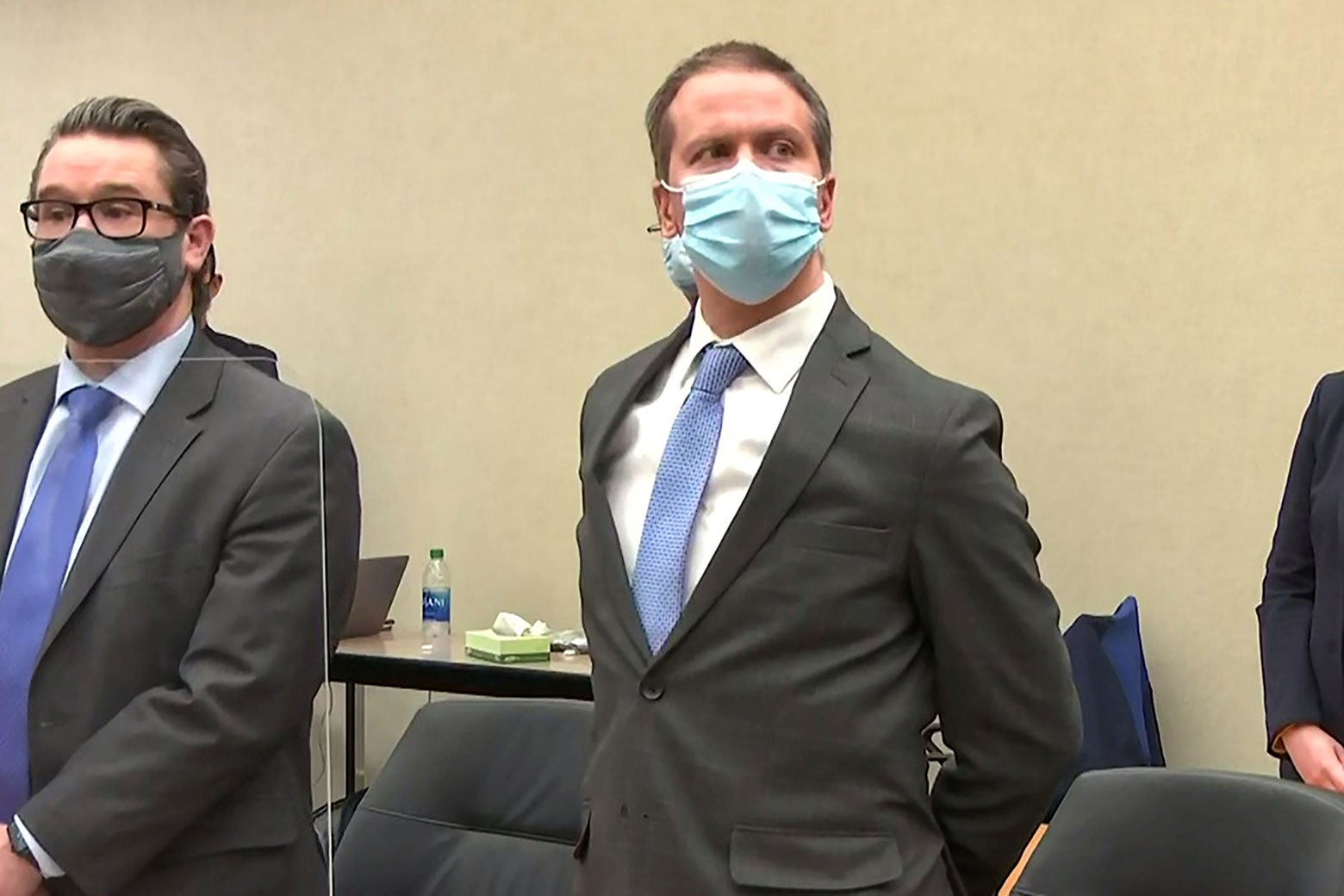 Derek Chauvin stands and looks toward the judge while wearing a mask and a suit with his hands behind his back