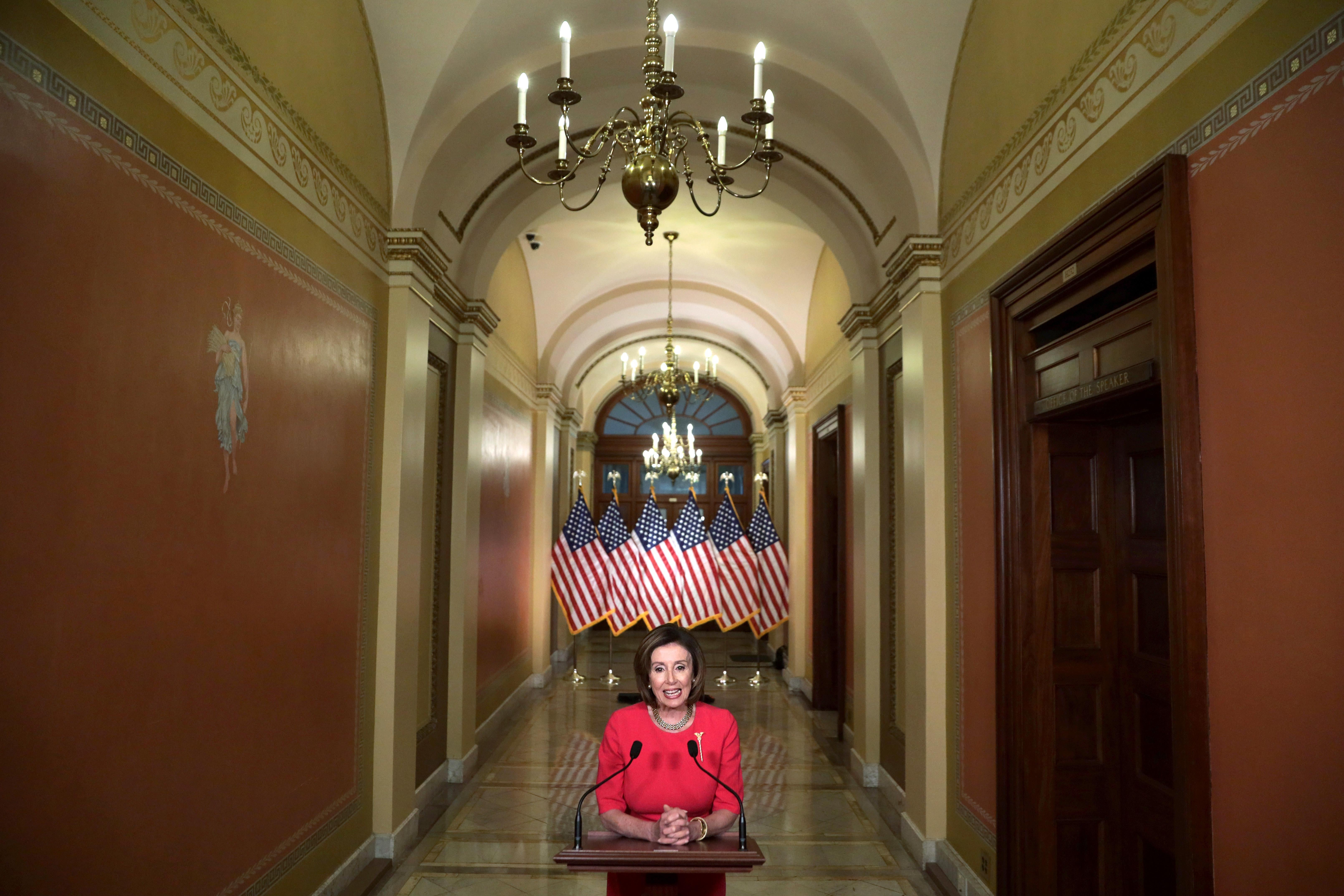 Nancy Pelosi stands at a lectern in a hallway. American flags stand at the back.