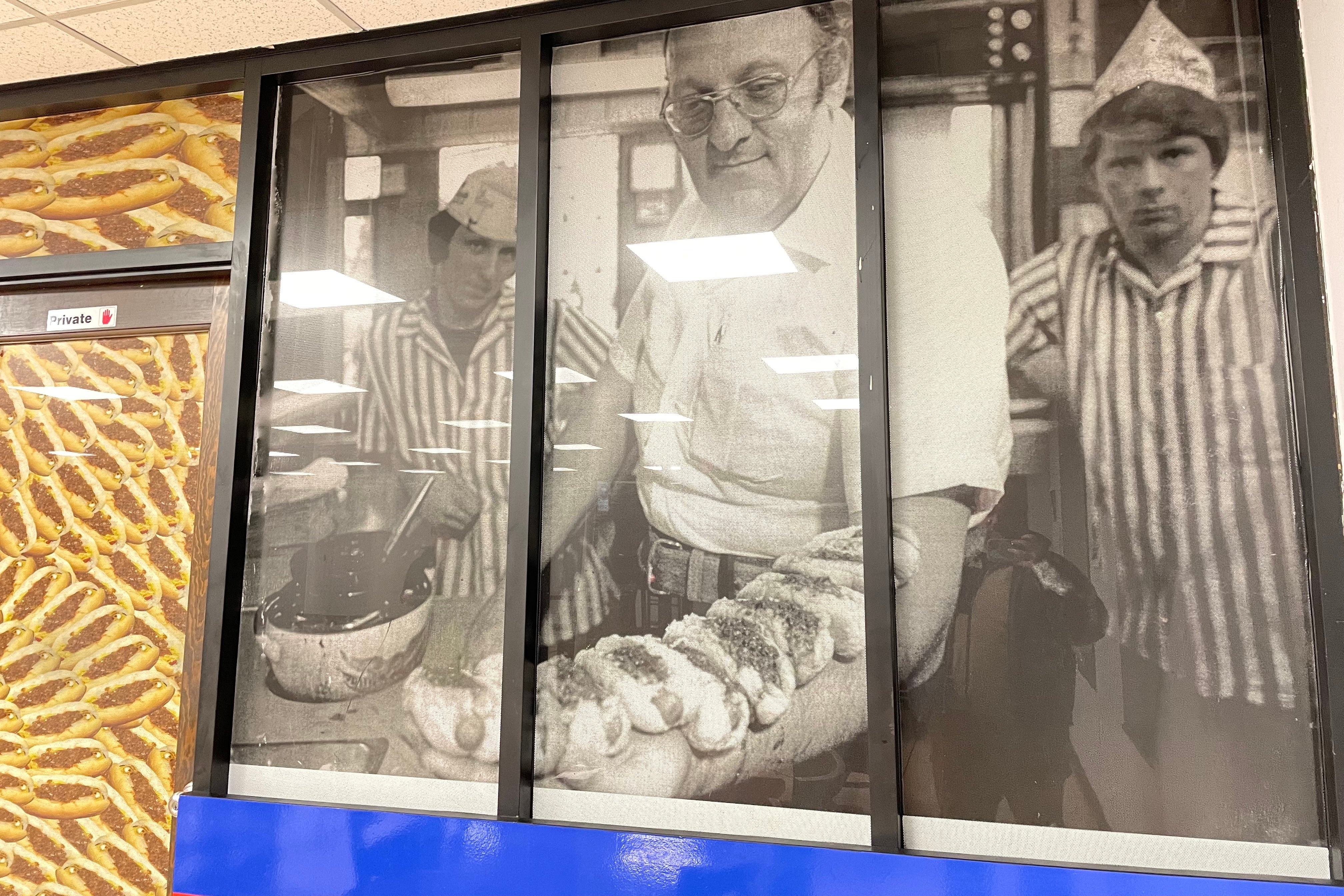 In an old black-and-white photo blown up on a restaurant wall, a man lines up mini hot dogs on his forearm.