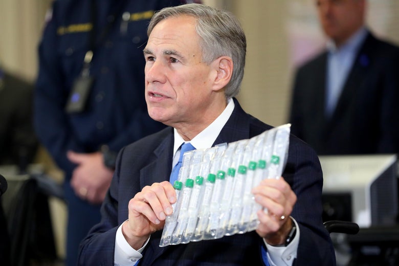 Greg Abbott, in a navy suit, holds up a clear plastic case containing several vials with green lids. Two men stand in the background.