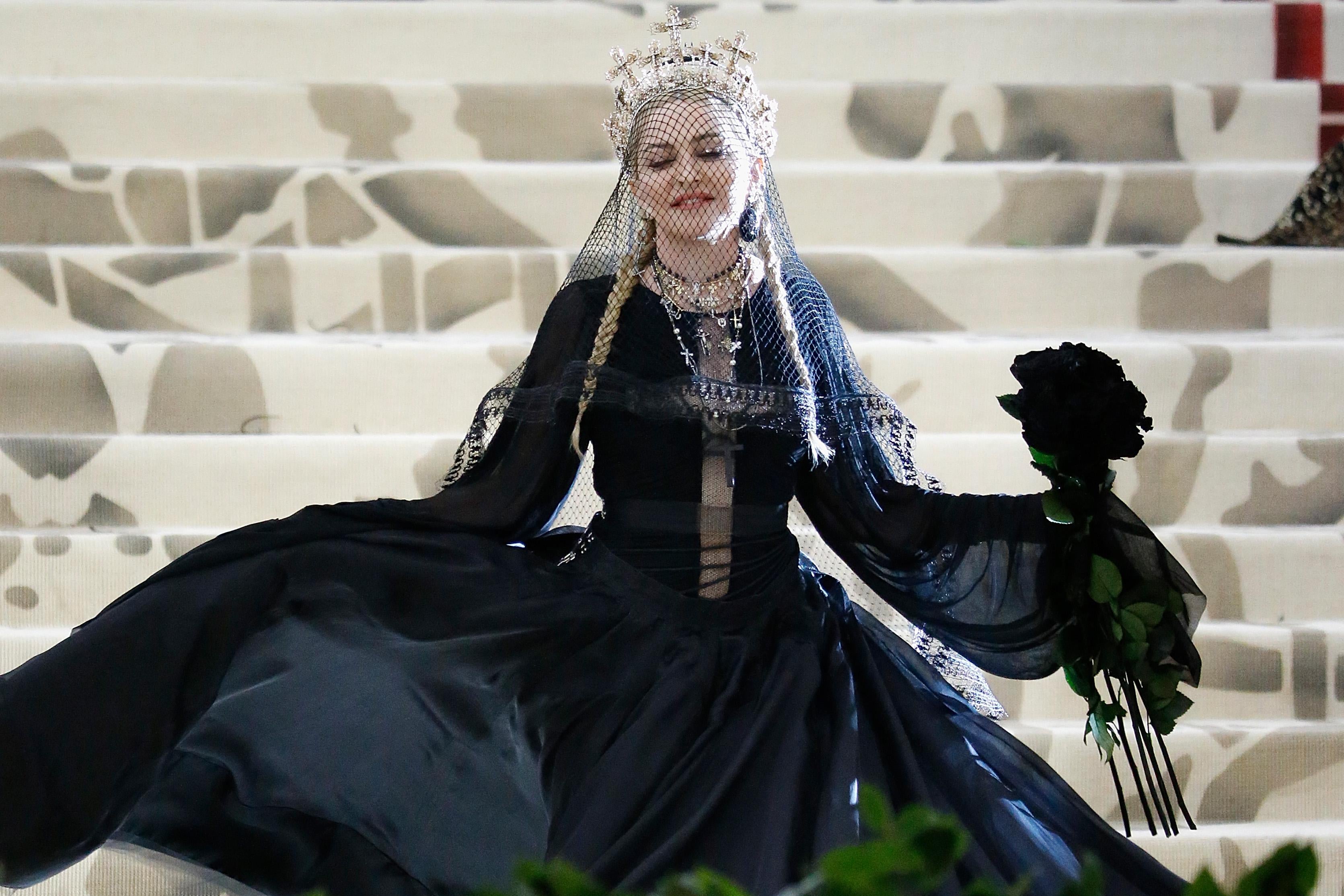 Madonna stands on the steps of the Met wearing a poofy black dress with multiple rosaries. She wears a crown made of crosses.