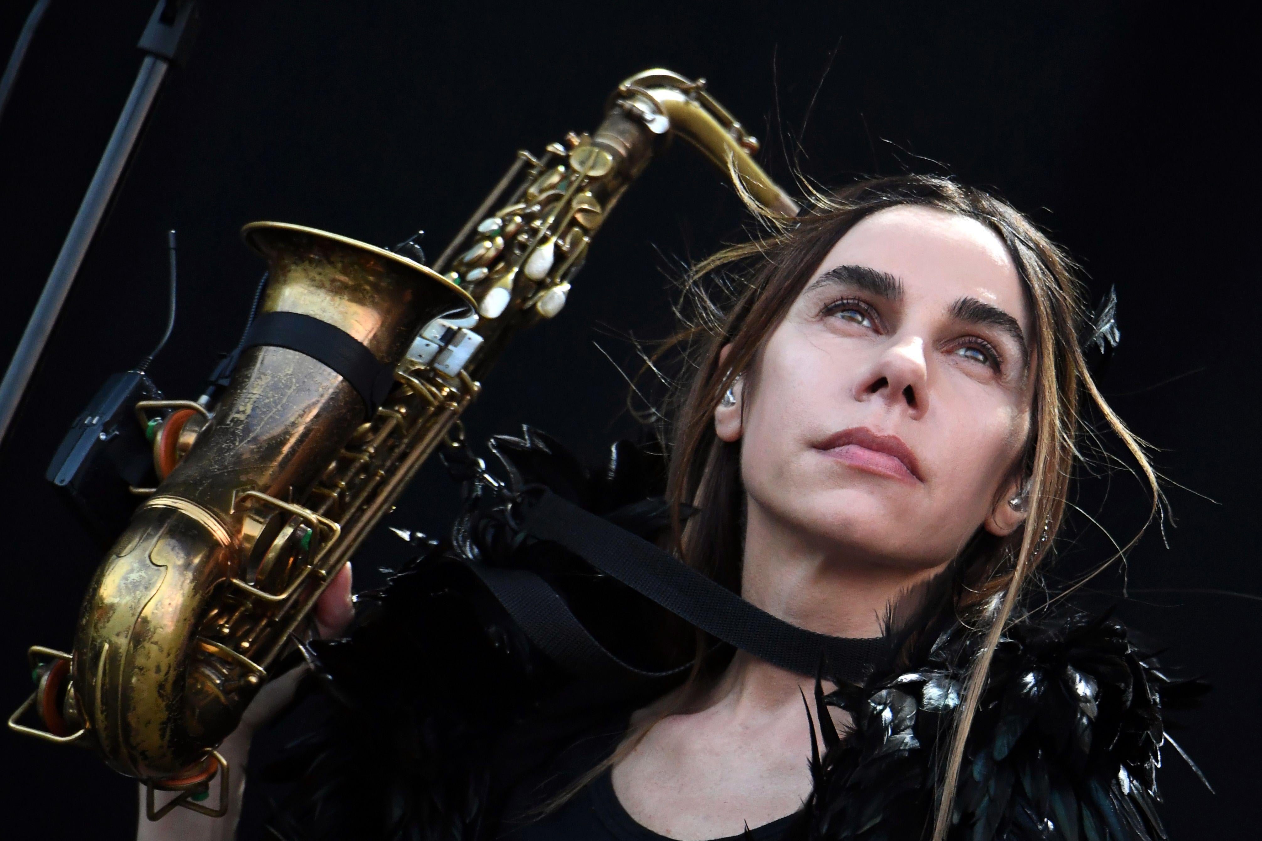 PJ Harvey, with an aloof stare and black-feathered get-up, holds a saxophone in front of a black background