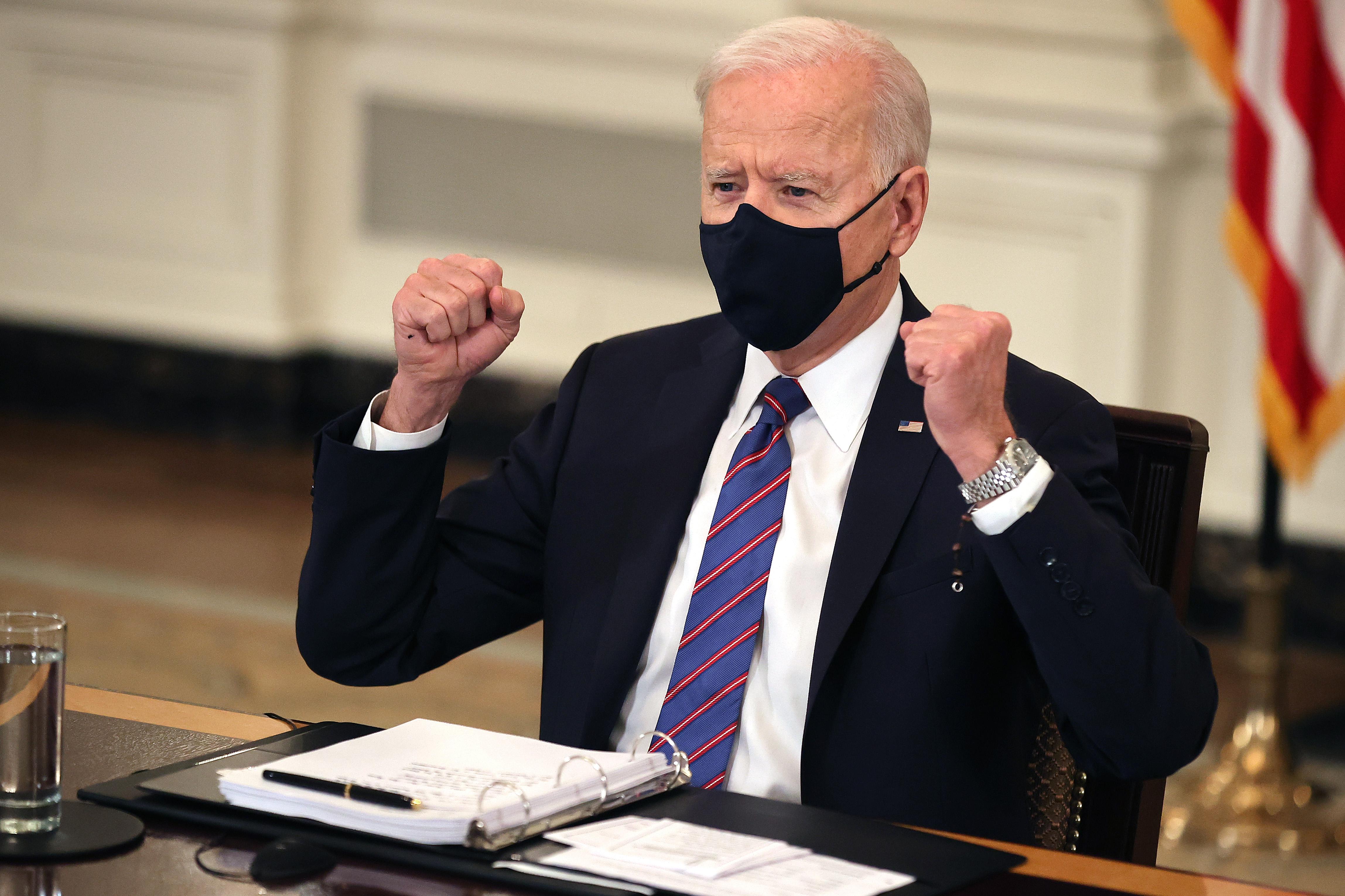 Joe Biden, wearing a face mask, sits at a table and raises his fists.