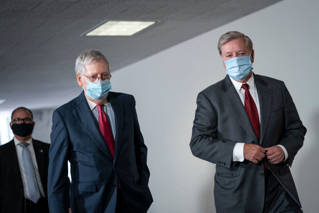 The two men, both wearing suits and blue masks, walk side by side in a hallway.