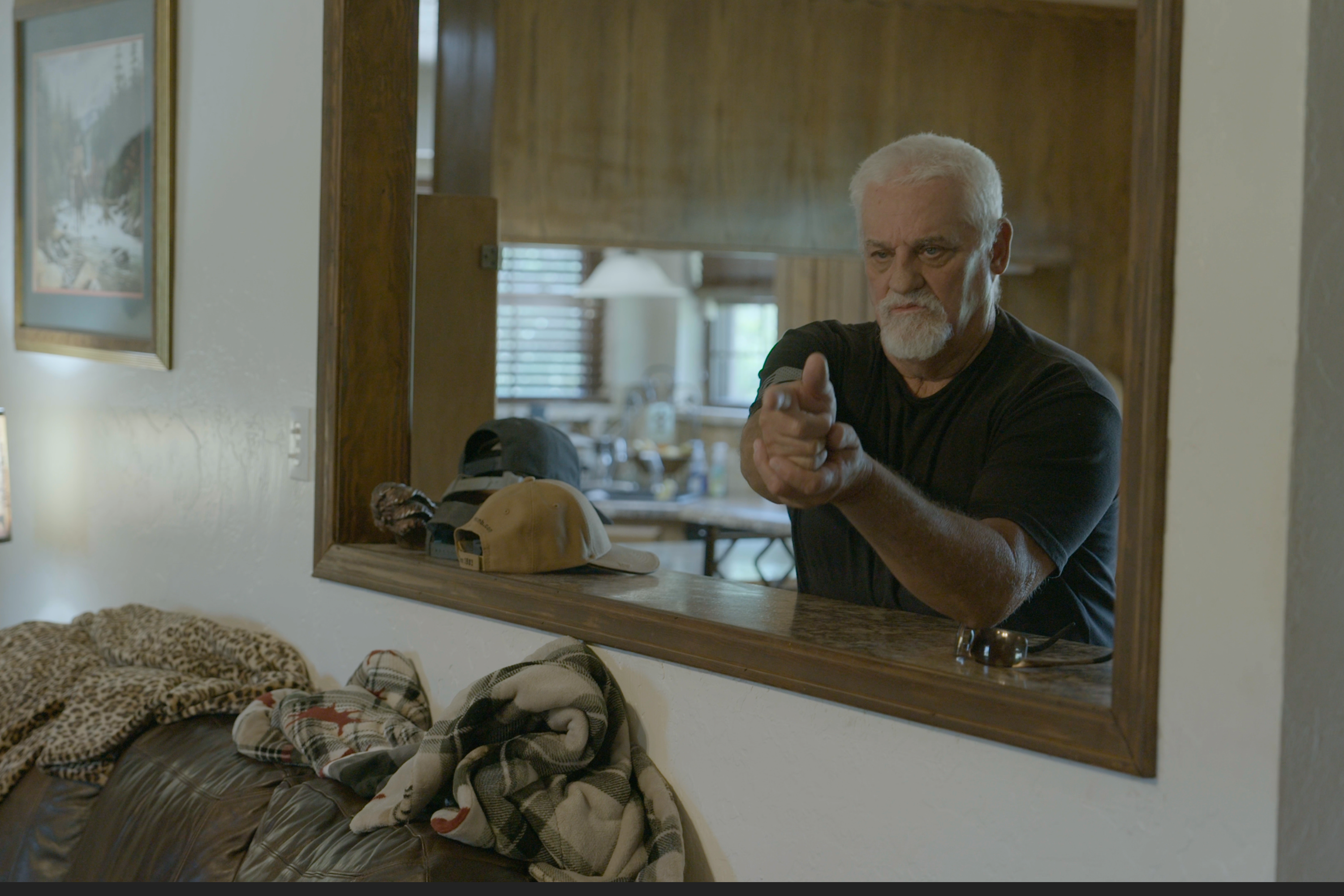 A silver-haired white man mimes pointing a gun while standing in a kitchen.