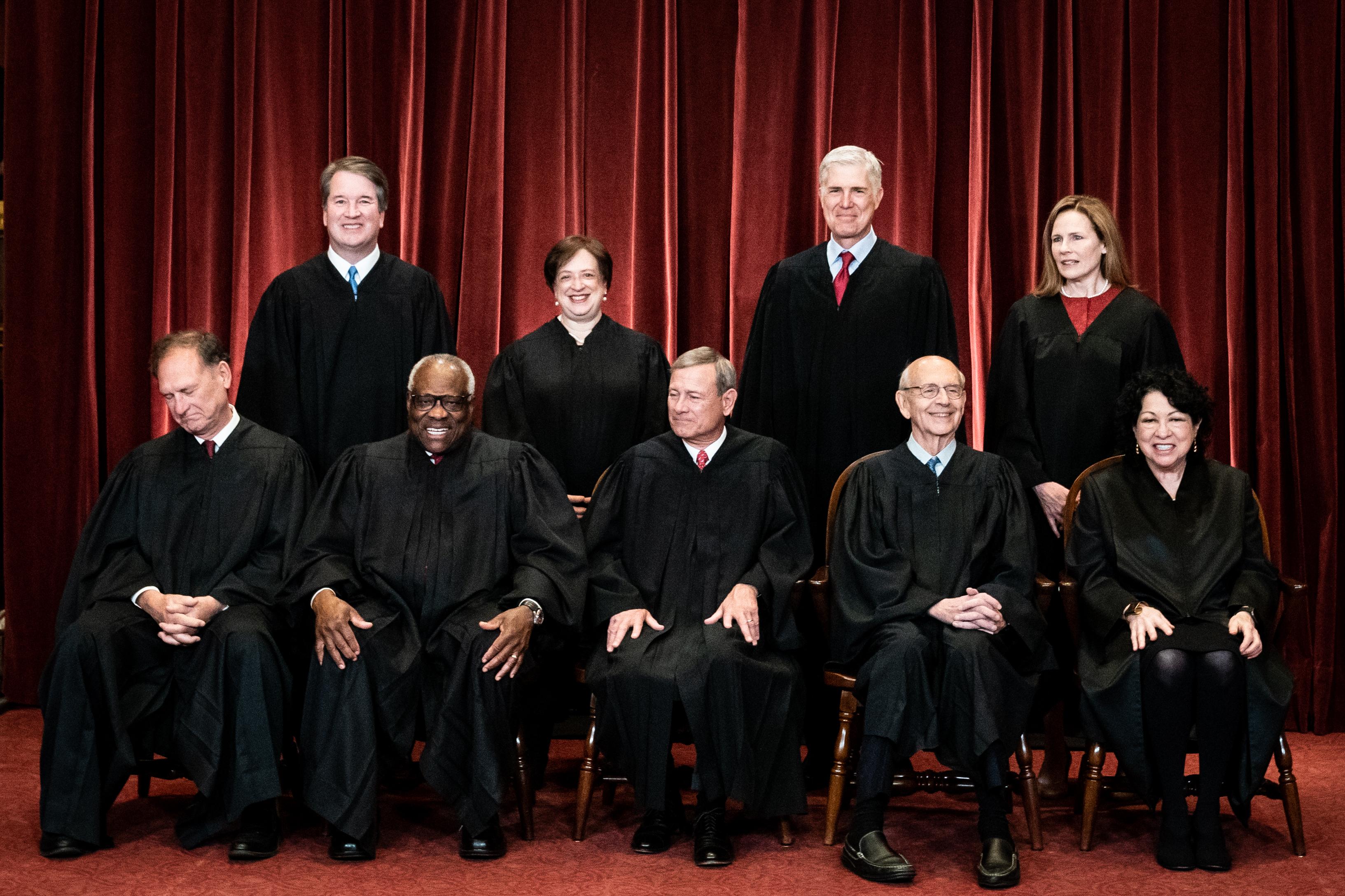 Candid photo of the justices laughing and smiling and seated in their robes in front of a red curtain