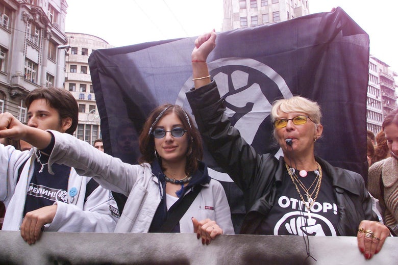 A young woman and her teacher hold up their fists in front of an "OTPOR" flag.