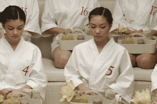 A woman, in a group of other women, wears a white robe embroidered with the number 5 and looks up at the camera.