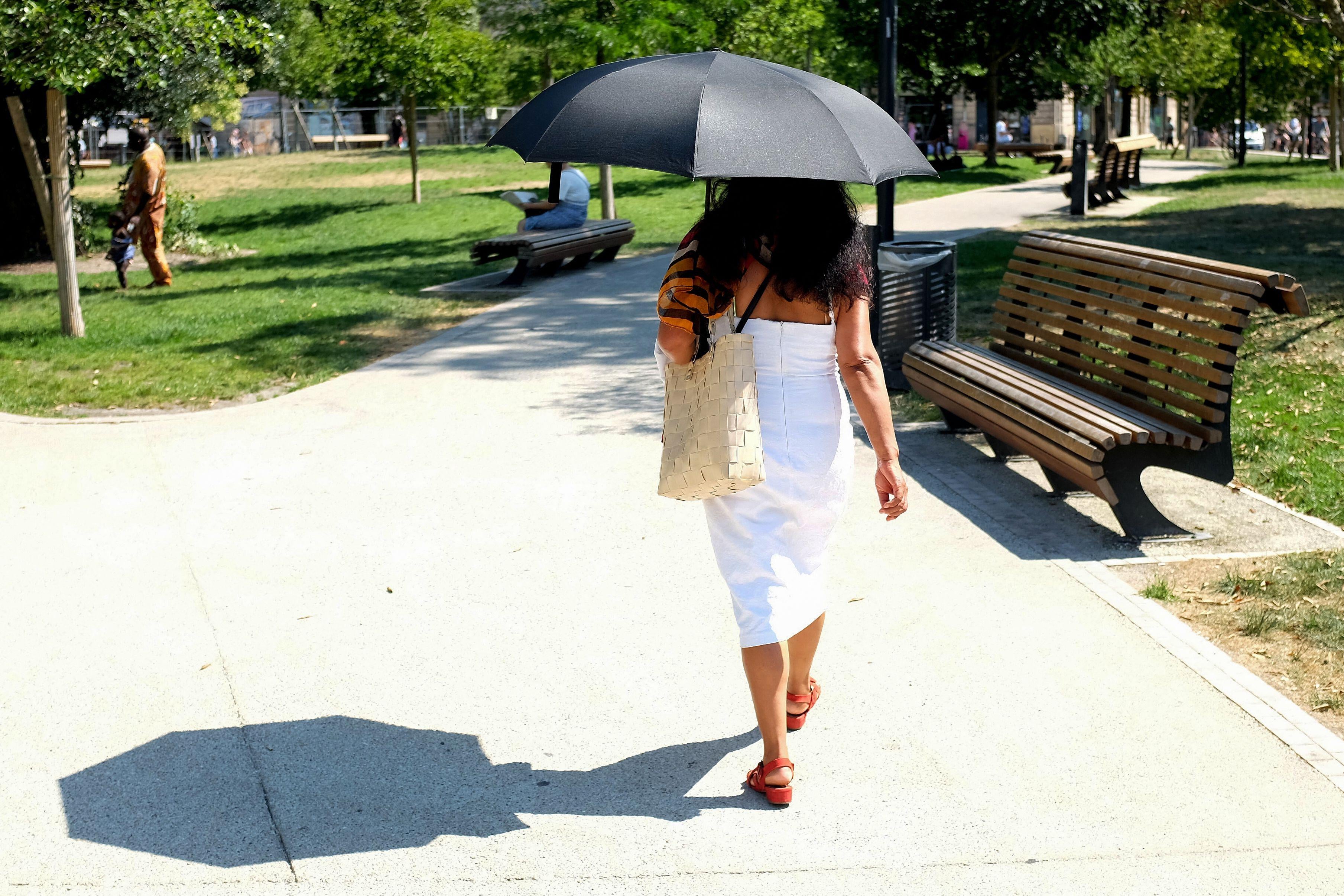 A woman walks in a park wearing a white dress and holding a black umbrella that casts a shadow on the sidewalk.