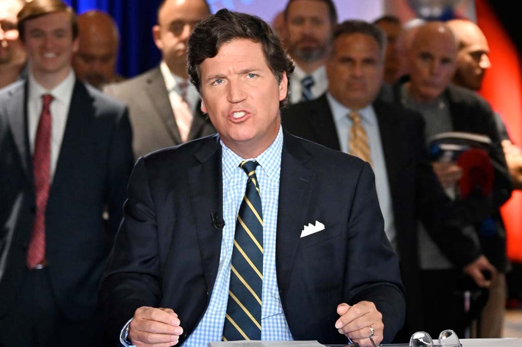 Carlson speaks toward a foregrounded camera from behind a desk on a television set. A number of audience members stand behind him.