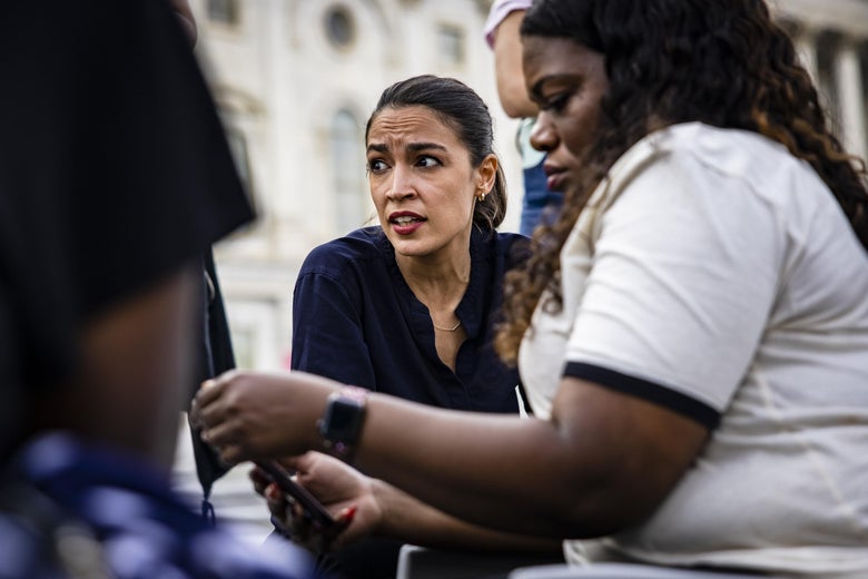 Ocasio-Cortez looks frustrated and Cori Bush looks at her phone as they sit on the steps