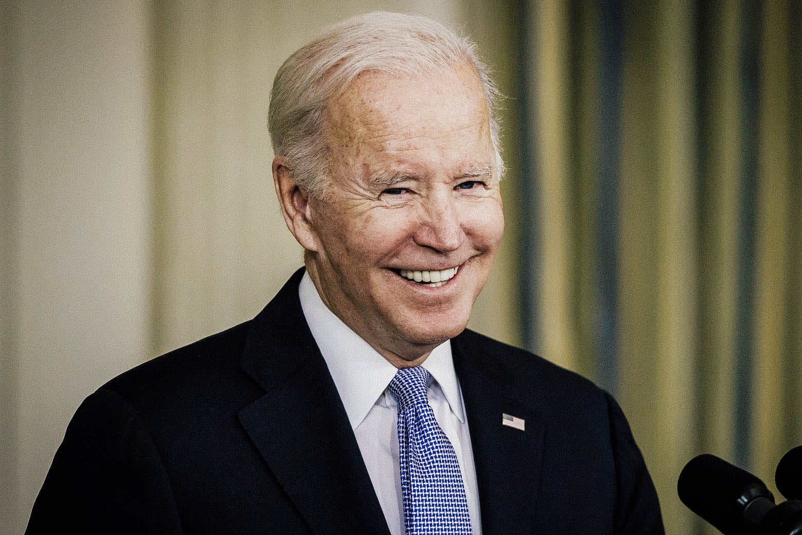 Biden smiles while standing at a podium behind a microphone.