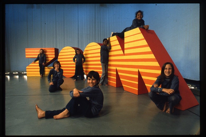 A group of barefoot kids sits on big orange-and-yellow-striped letters that read: ZOOM. 
