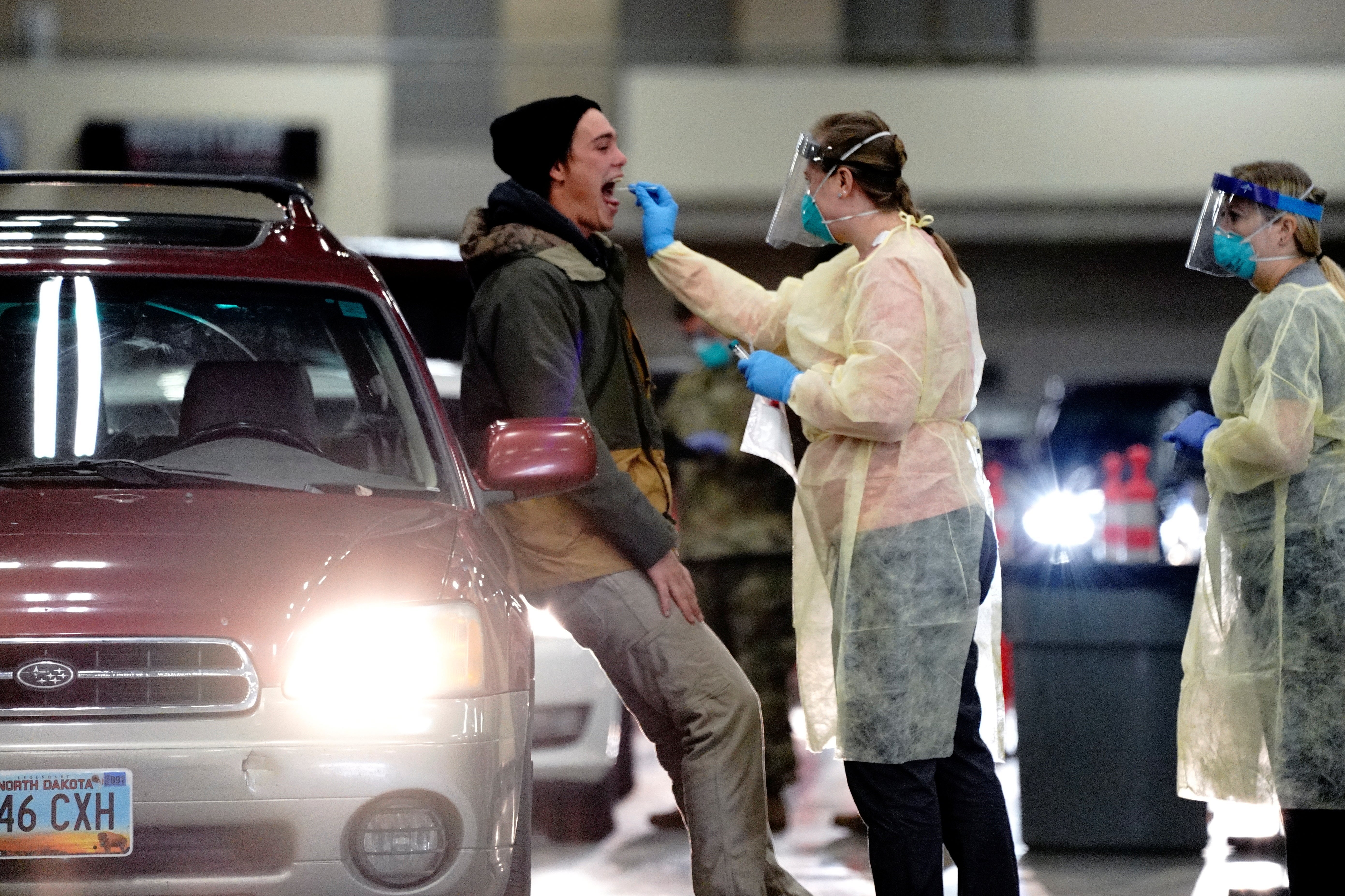 A medical worker in PPE swaps a man's throat as he stands beside a car.