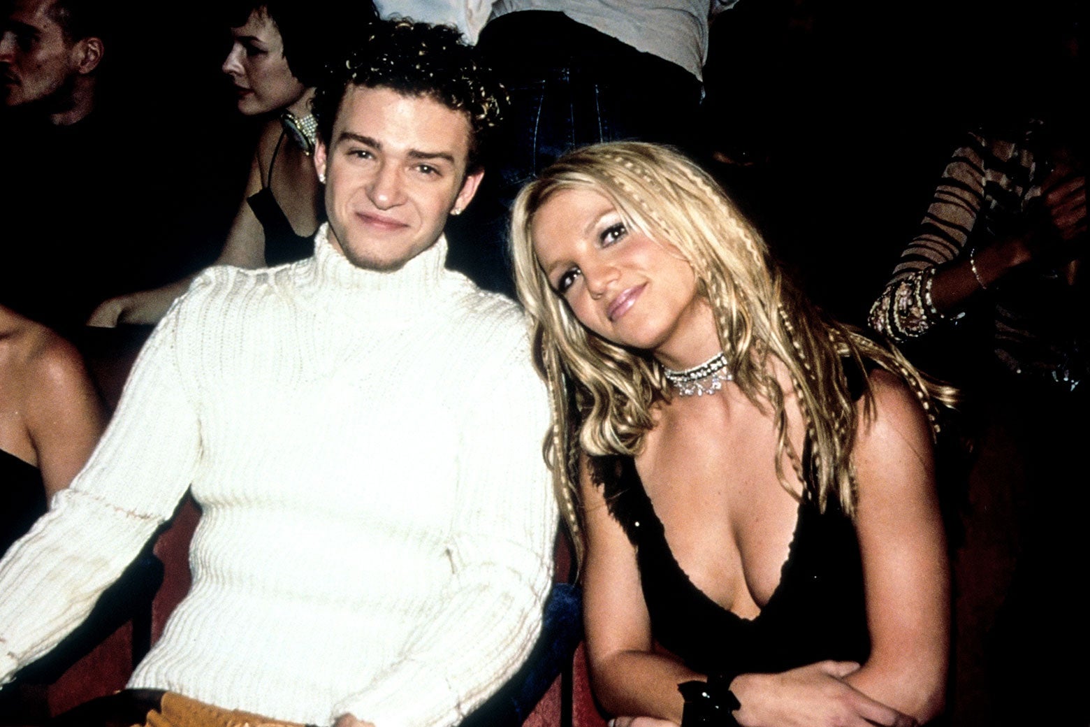 Justin in a white turtleneck sweater and Britney in a low-cut black top lean toward each other while seated in the audience.