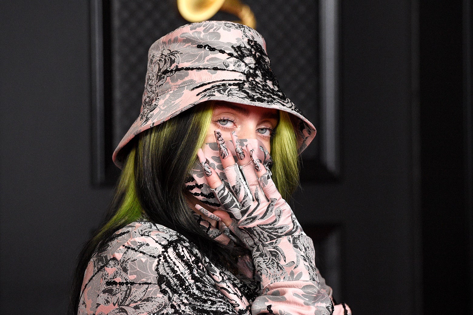 A girl with black-and-green hair wears a bucket hat with a pink, gray, and black floral-style pattern and a matching long-sleeve shirt. Her hand is covering her face as she eyes the camera. 