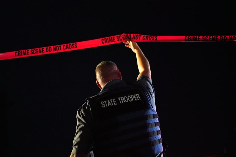 A man in a state trooper vest holds up crime scene tape