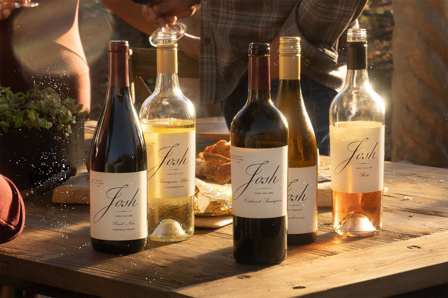 Multiple bottles of Josh wine sit on a rustic-looking wooden table, with the sunlight streaming behind them.