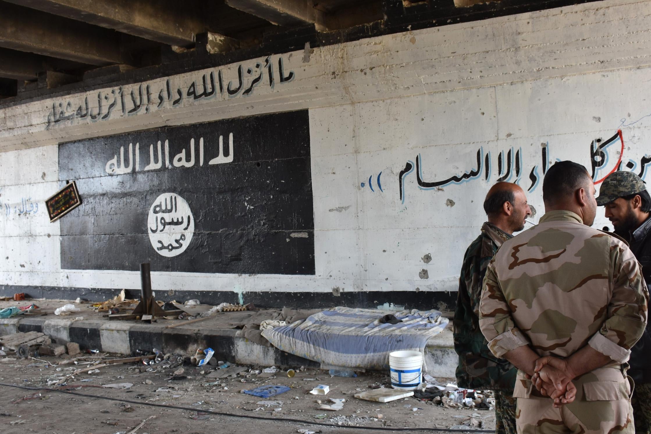 Men stand talking in a tunnel in front of a wall with graffiti of the ISIS flag.