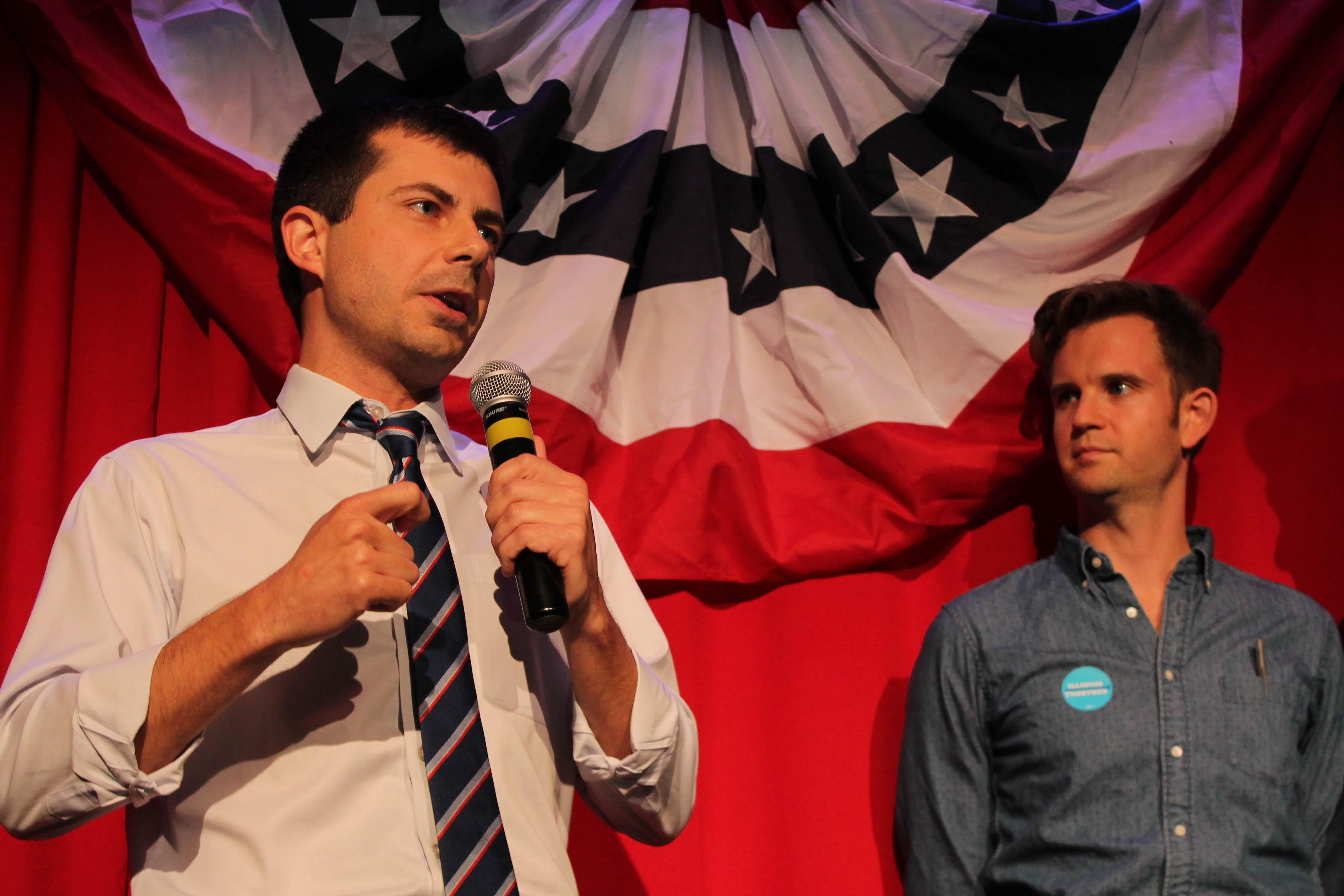 Standing in front of a patriotic-looking background, Buttigieg speaks into a microphone.