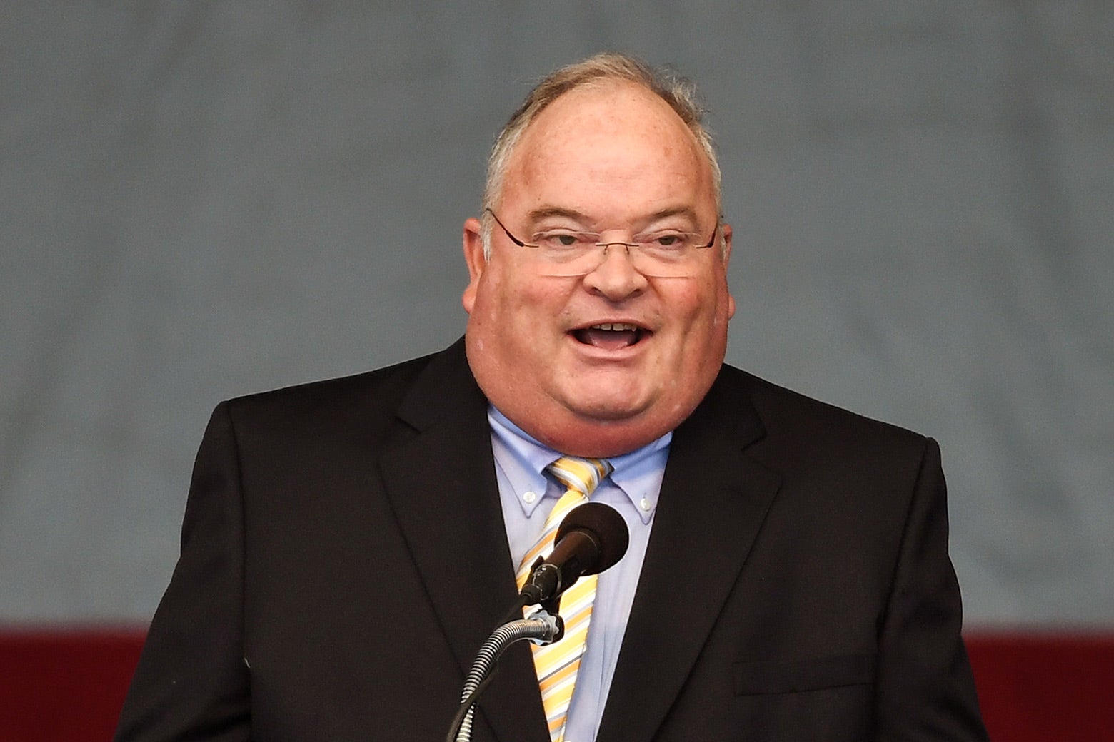 A man in a suit and with a very round face speaks into a microphone.