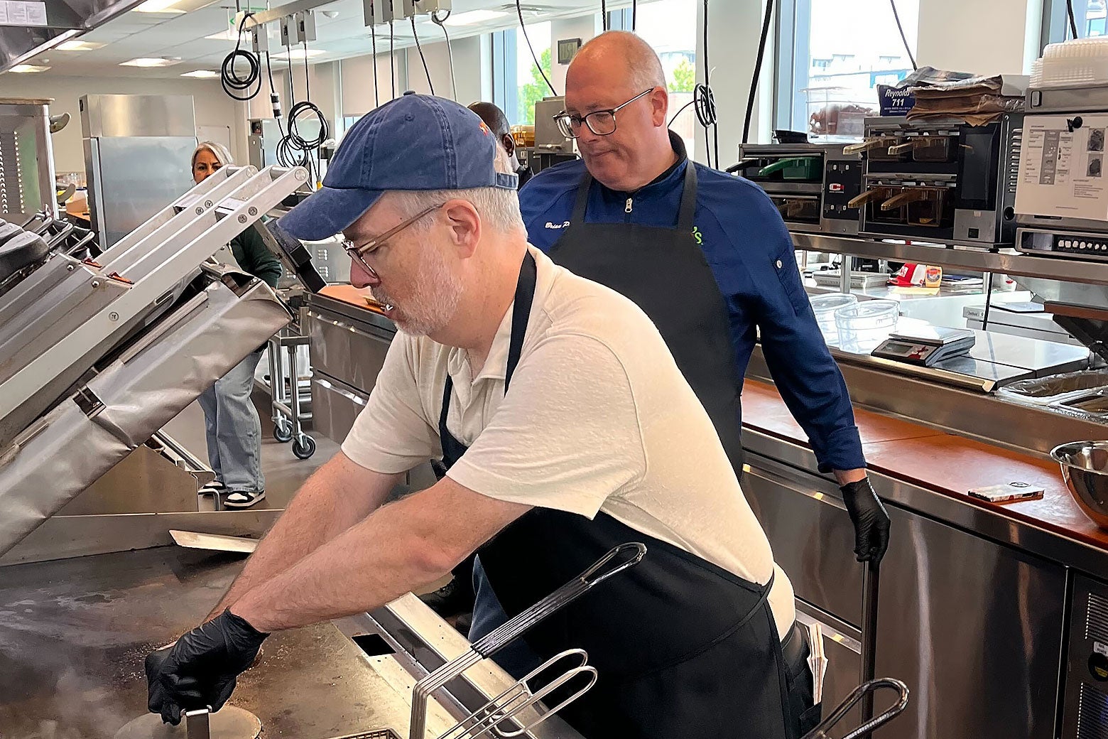 Two men inside a kitchen, with one cooking a burger.
