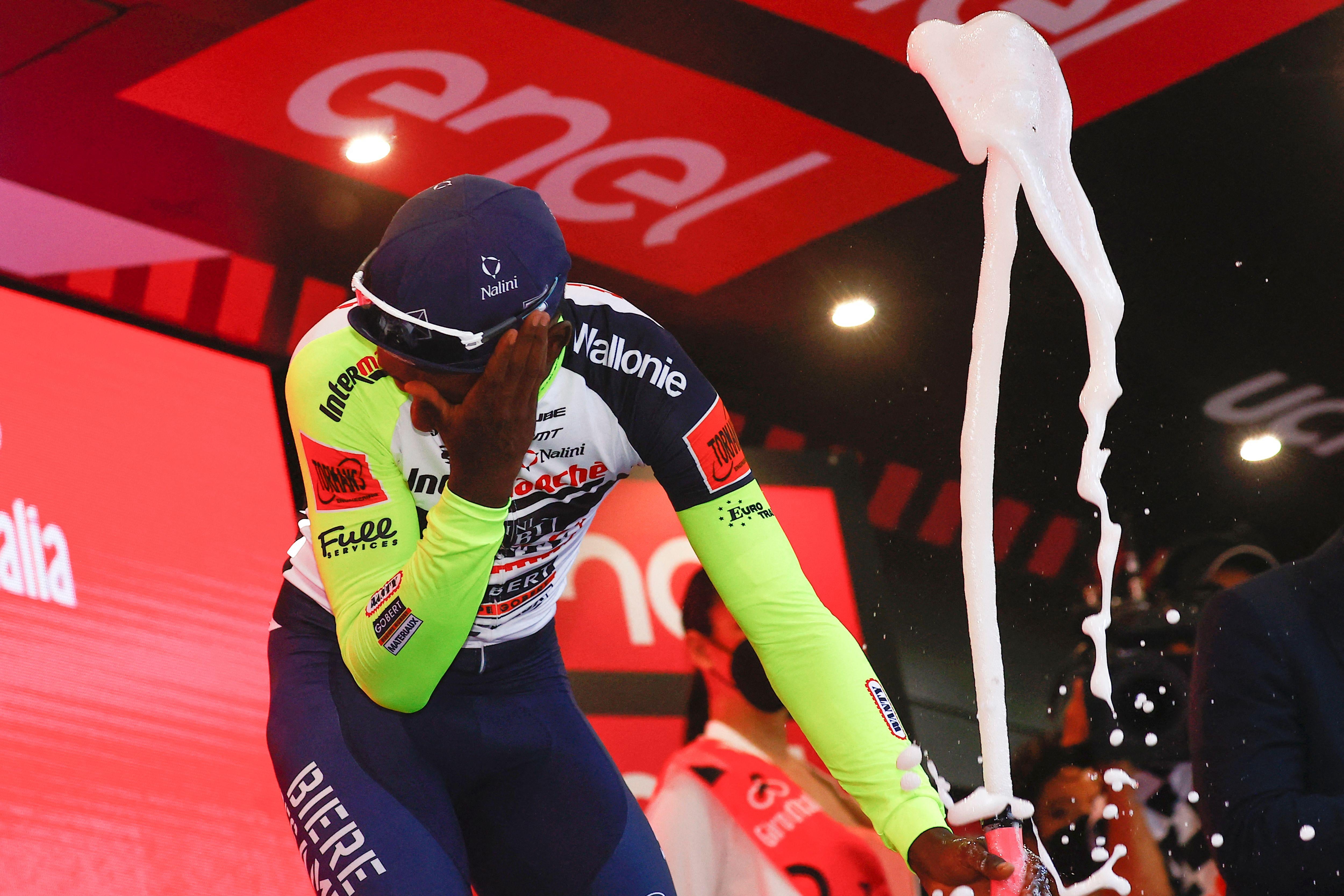 A cyclist winces and holds a hand up to his left eye as he holds a spewing Prosecco bottle in his other hand.