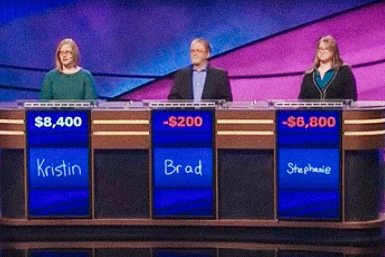 Three Jeopardy! contestants stand at their tables, with their scores and names displayed below.