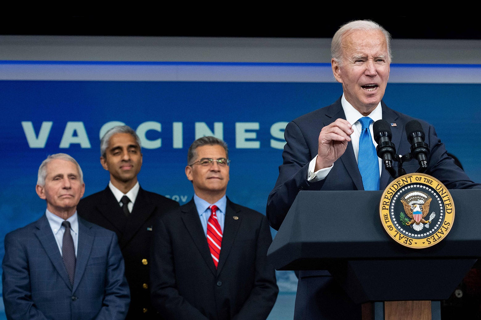 Joe Biden speaking at a podium with POTUS seal as three administration officials look on behind him.