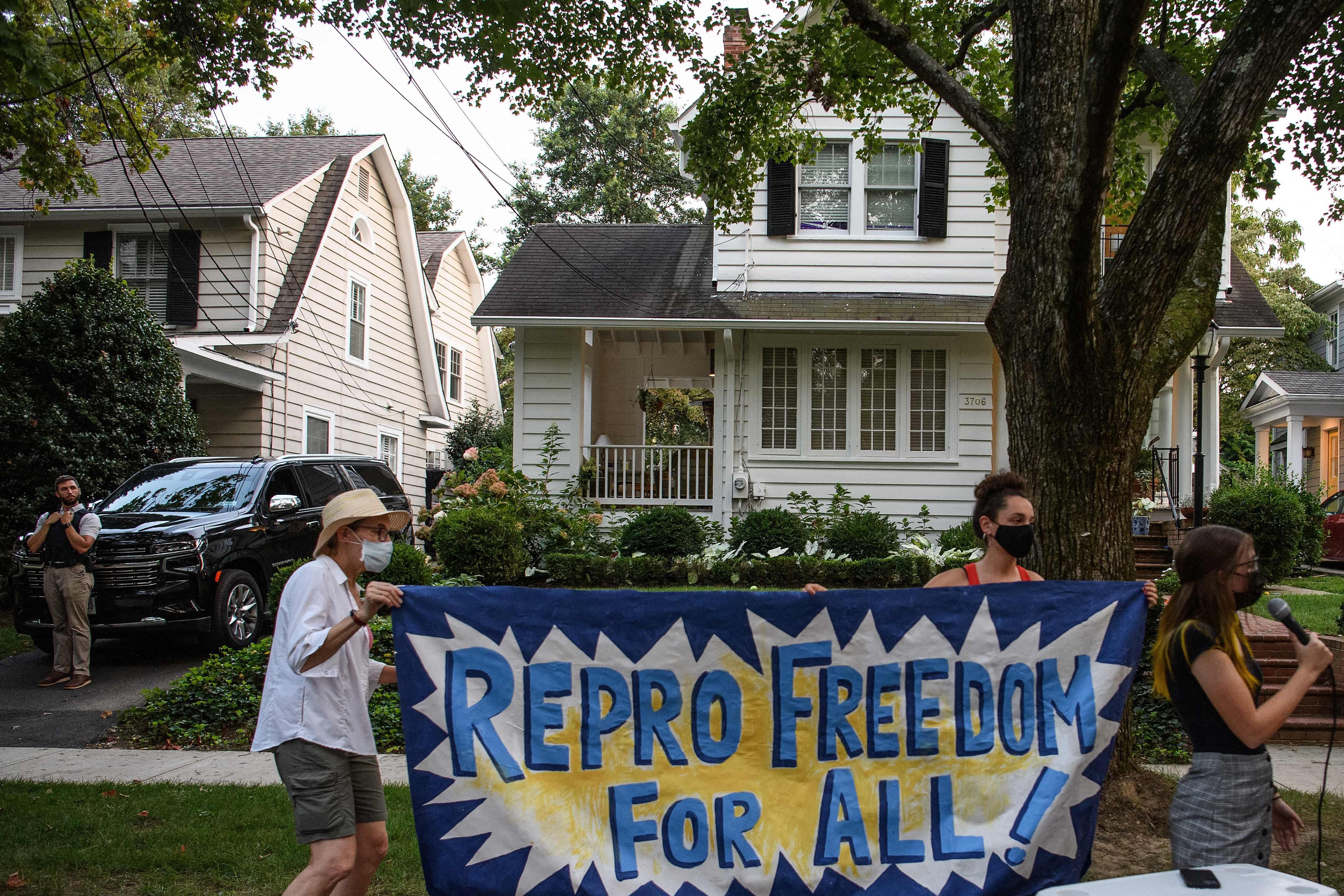 Two people hold up a sign that says "Repro Freedom for All!" as another person speaks into a mic on a suburban street