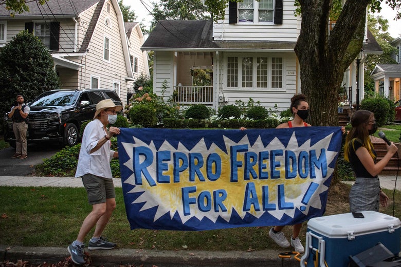 Two people hold up a sign that says "Repro Freedom for All!" as another person speaks into a mic on a suburban street